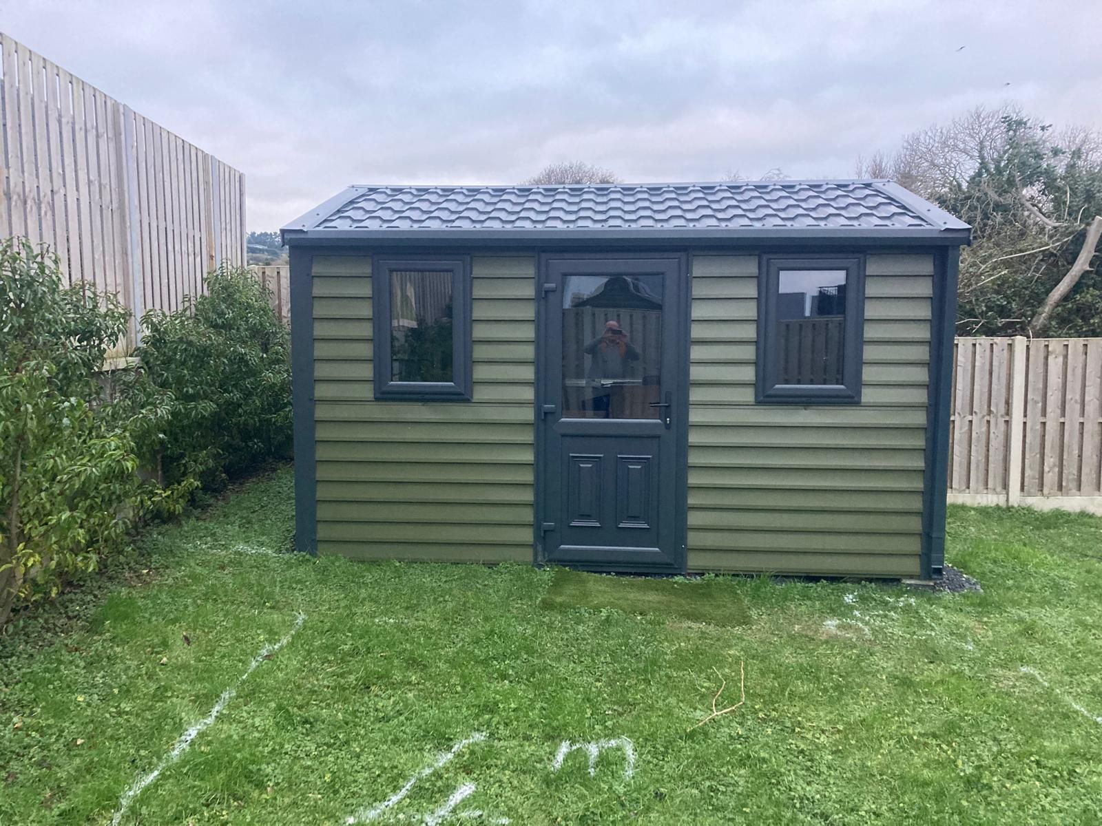 Green shed with dark gray trim, windows, and door in a grassy yard, partially fenced.