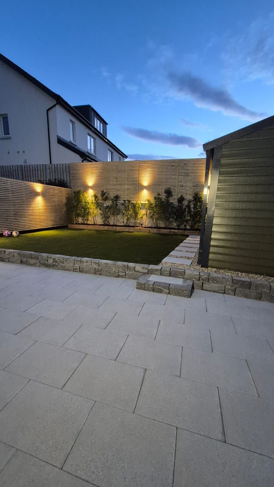Patio with stone pavers, grass, hedges, and uplighting at dusk. Wooden fence, shed, and houses in the background.