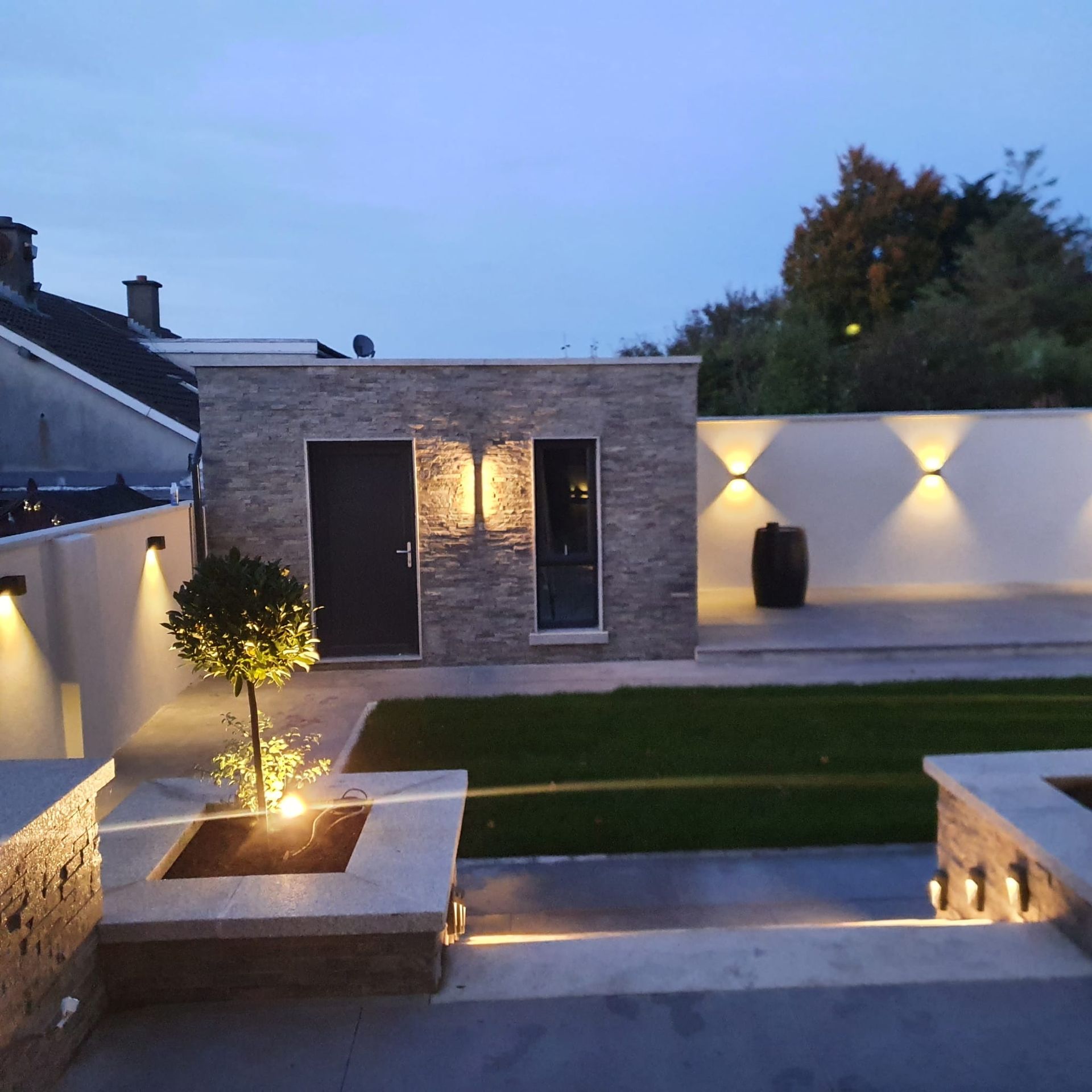Backyard patio with lit walls and building. Includes grass, small tree, and a barrel. Evening scene.