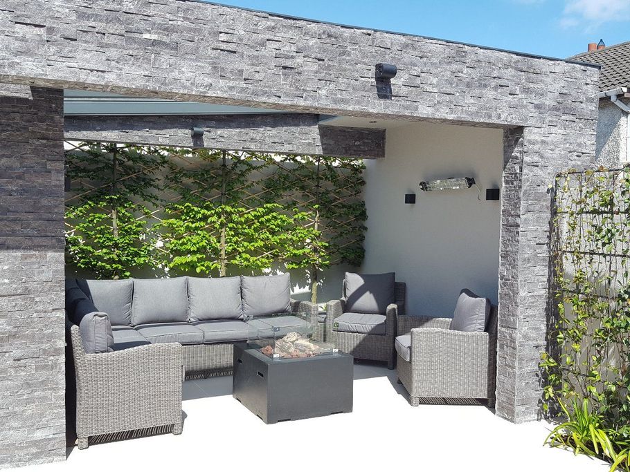 Outdoor seating area with gray stone walls, sofa, chairs, and fire pit. Green plants and vines.
