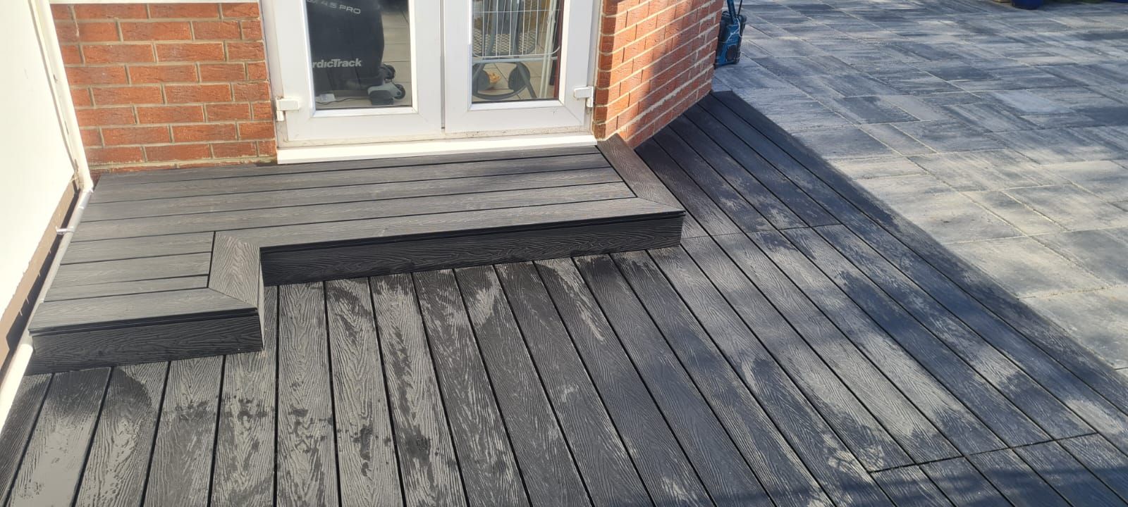 Composite decking with steps leading to a doorway, alongside brick exterior and paved patio.