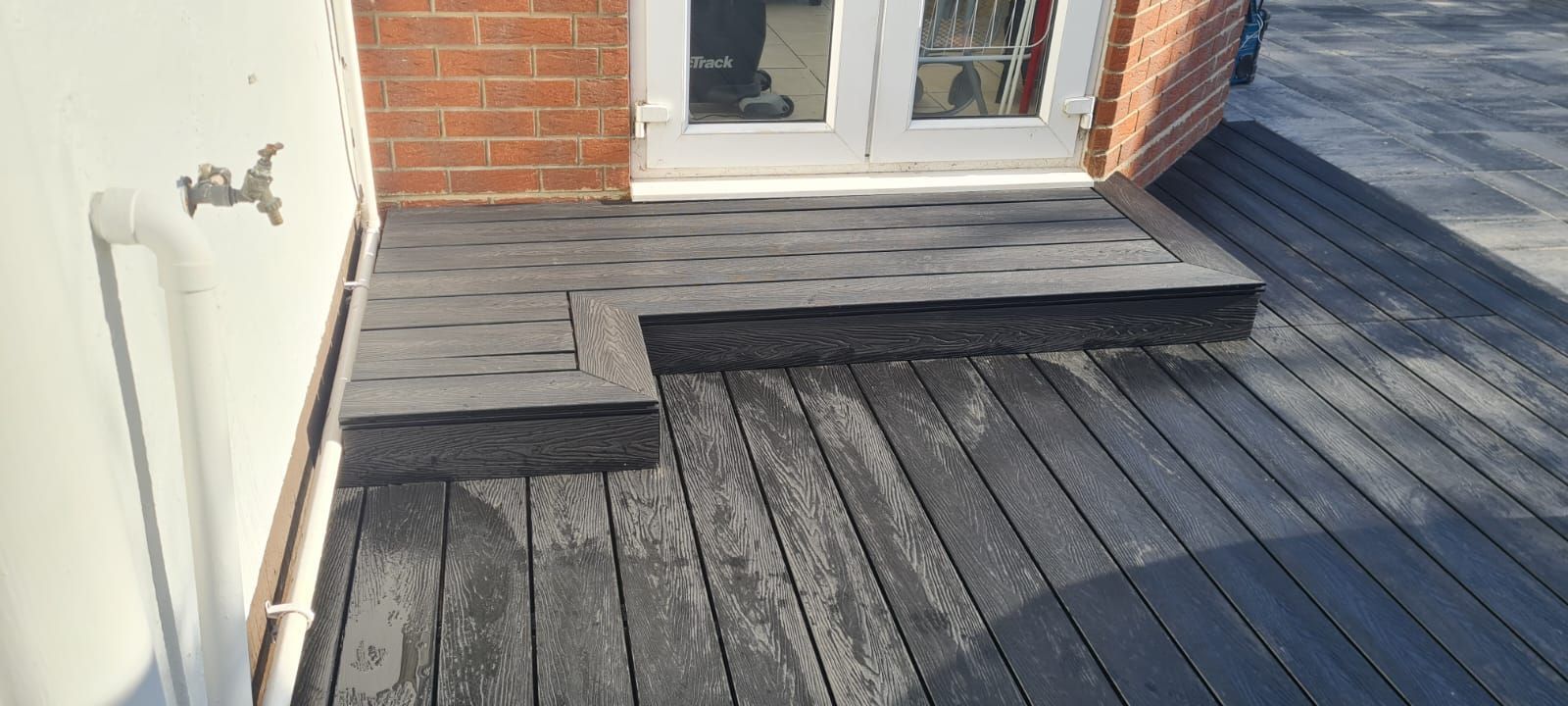 Composite decking leading to a set of doors. The decking has a hatch and a step up. A white wall is to the left.