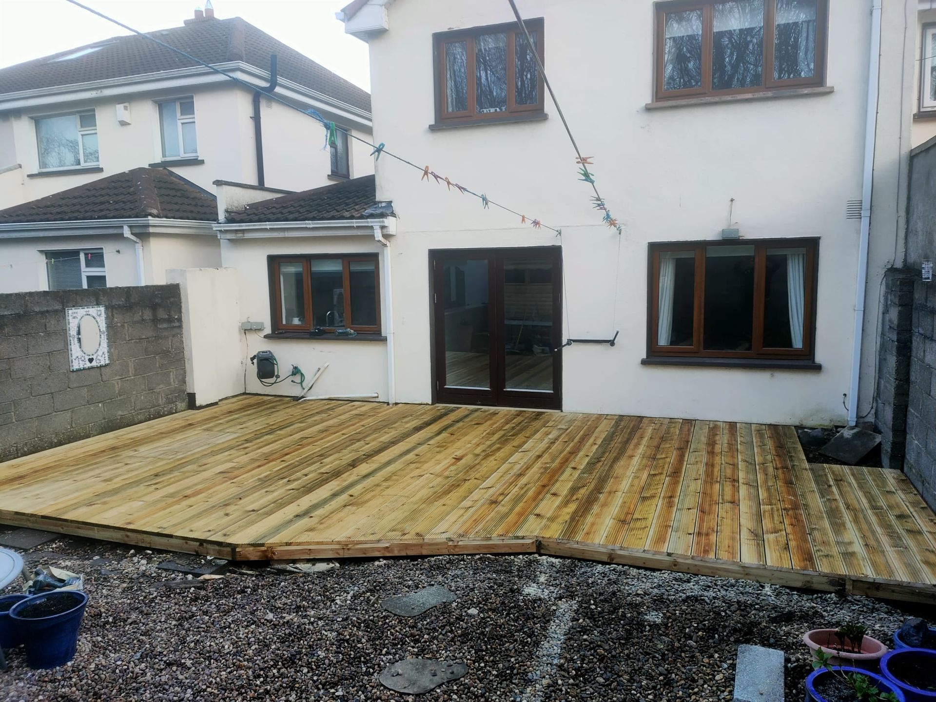 Wooden deck in a backyard, spanning the width of the house with two windows and a door. Gravel foreground.