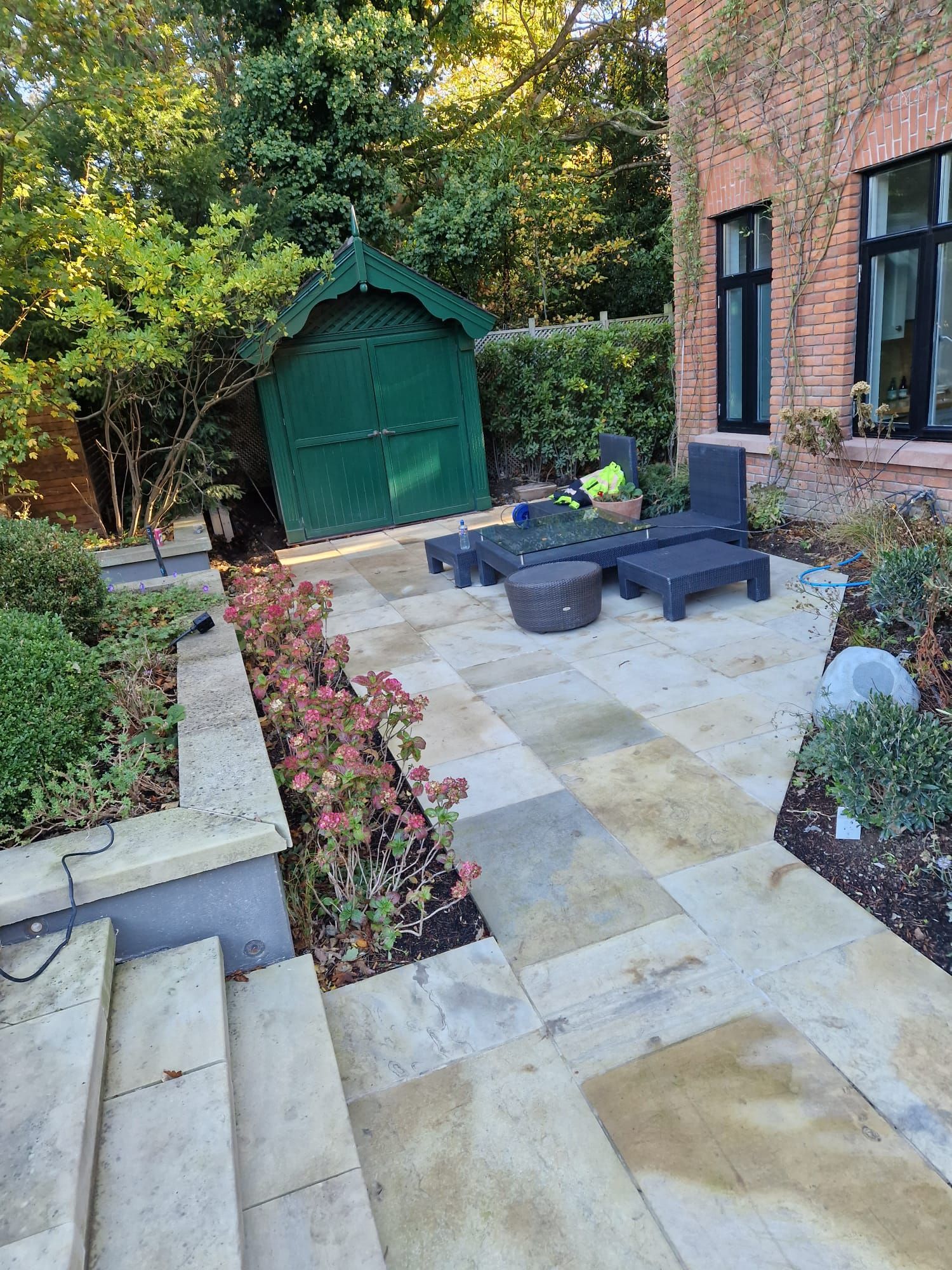 Stone patio with lounge chairs, shed, and flower beds near a brick building.