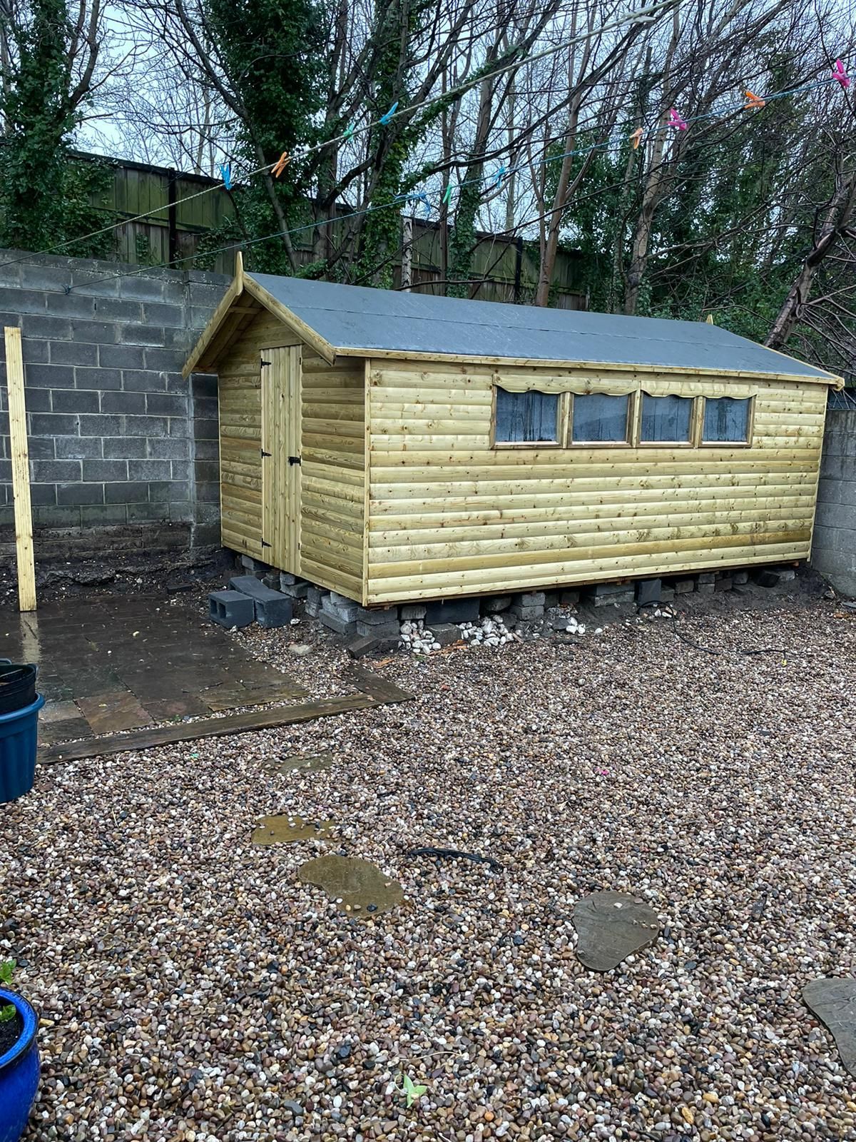 Wooden shed with a dark gray roof sits on concrete blocks in a gravel yard.