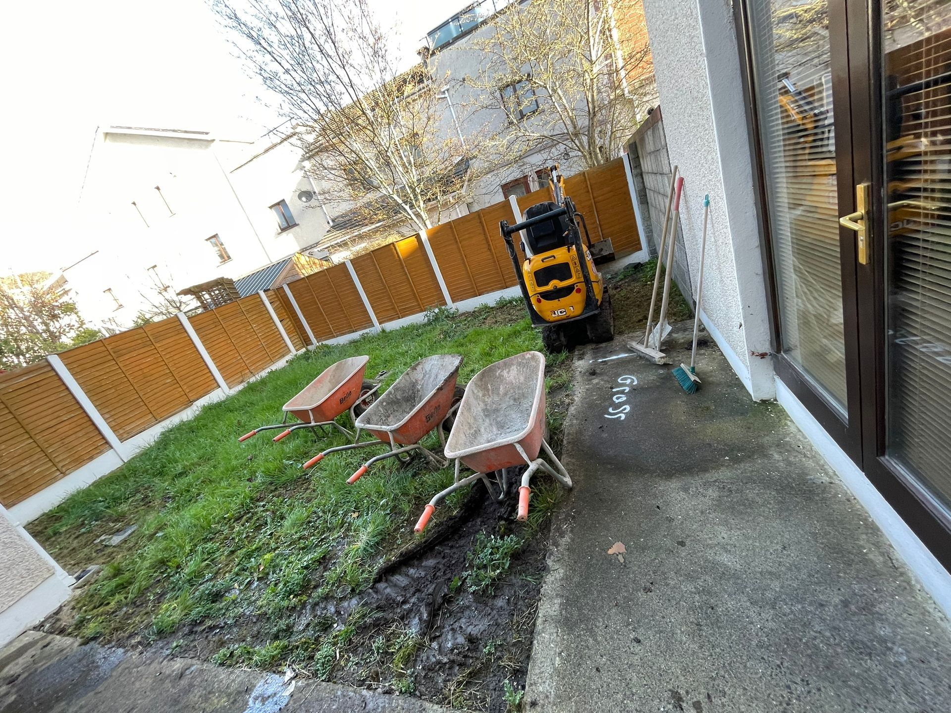 Lawn with three wheelbarrows, small excavator, brown fence, and concrete patio by a house door.