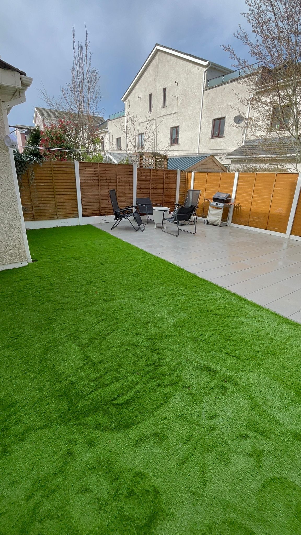 Backyard with artificial green grass, concrete patio, and wooden fence. Patio furniture and grill sit on the patio.