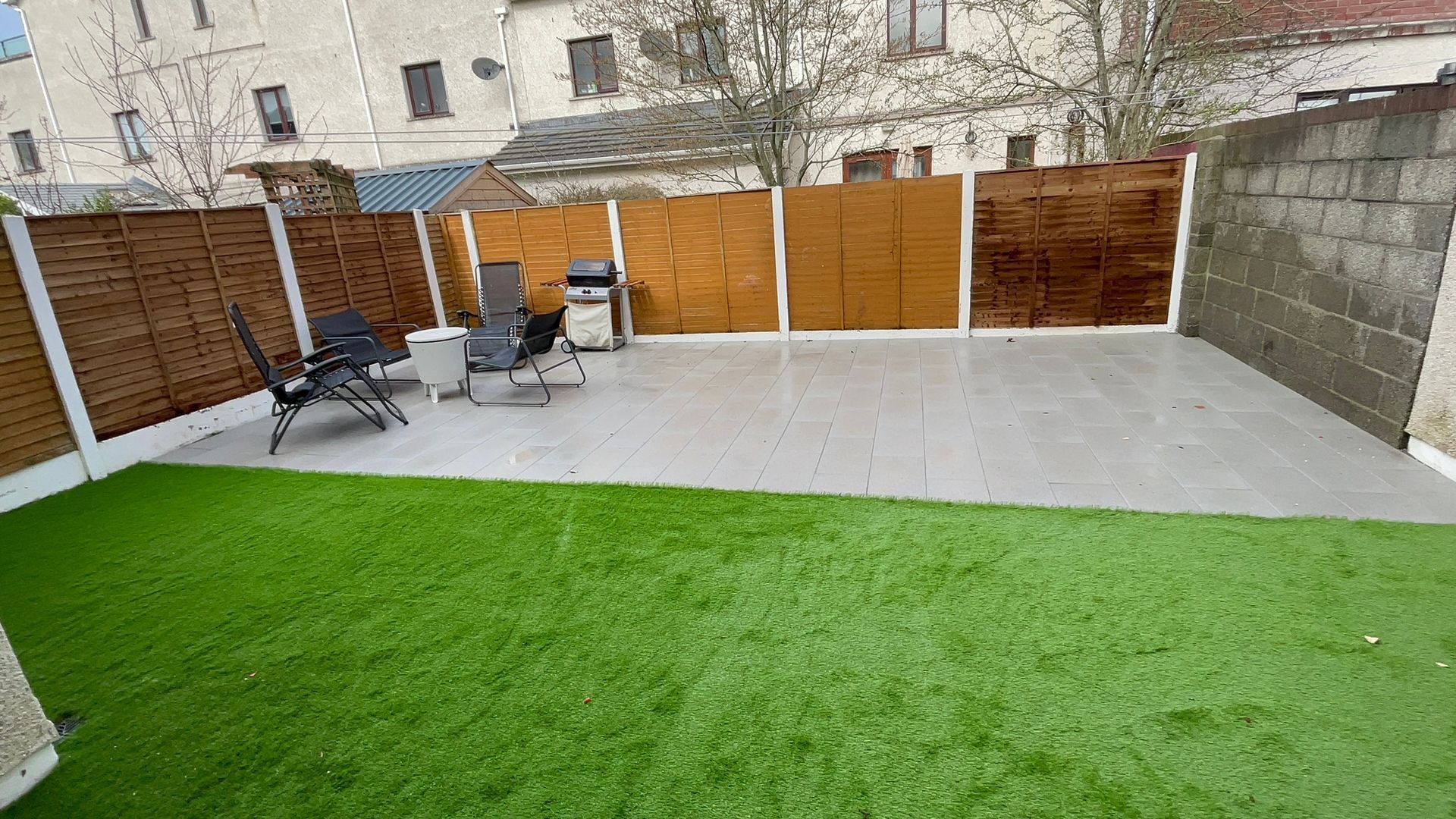 Backyard with artificial grass and patio; wooden fence, grill, chairs.