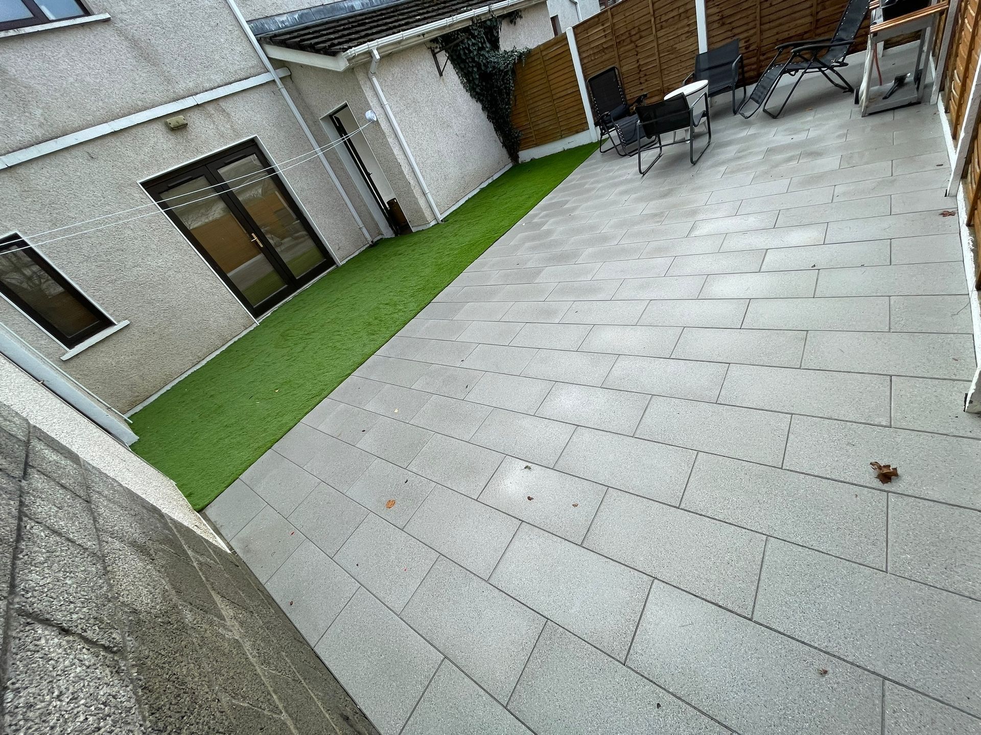 Backyard patio with gray pavers and green turf strip. Black chairs sit on the patio.