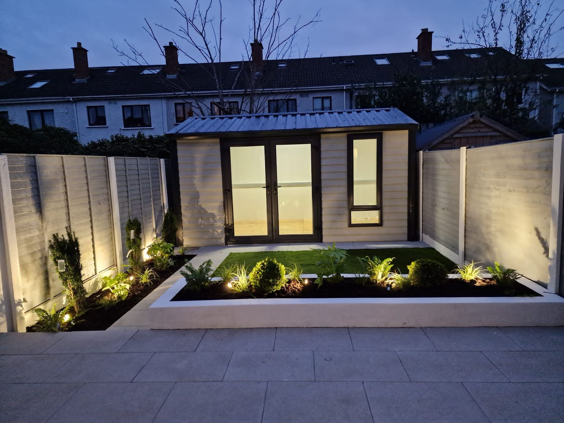 Backyard shed with lighting, surrounded by a garden bed and concrete fencing at dusk.