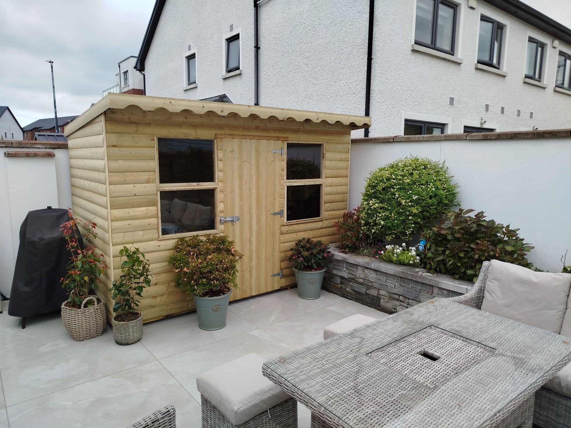 Wooden shed in a patio area with outdoor seating and plants, next to a white building.