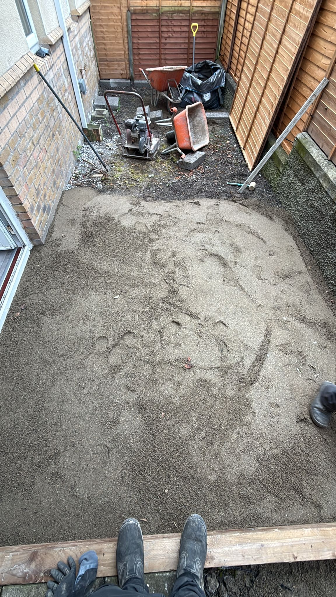 Overhead view of a small backyard covered in gravel and dirt; construction debris and wheelbarrow in the background.