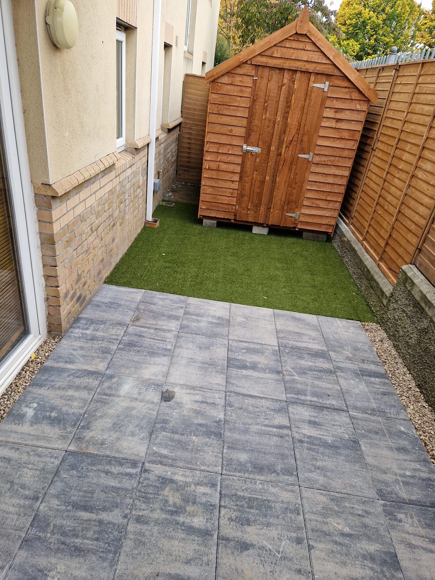 Patio with gray pavers, artificial grass, and a wooden shed against a building and fence.