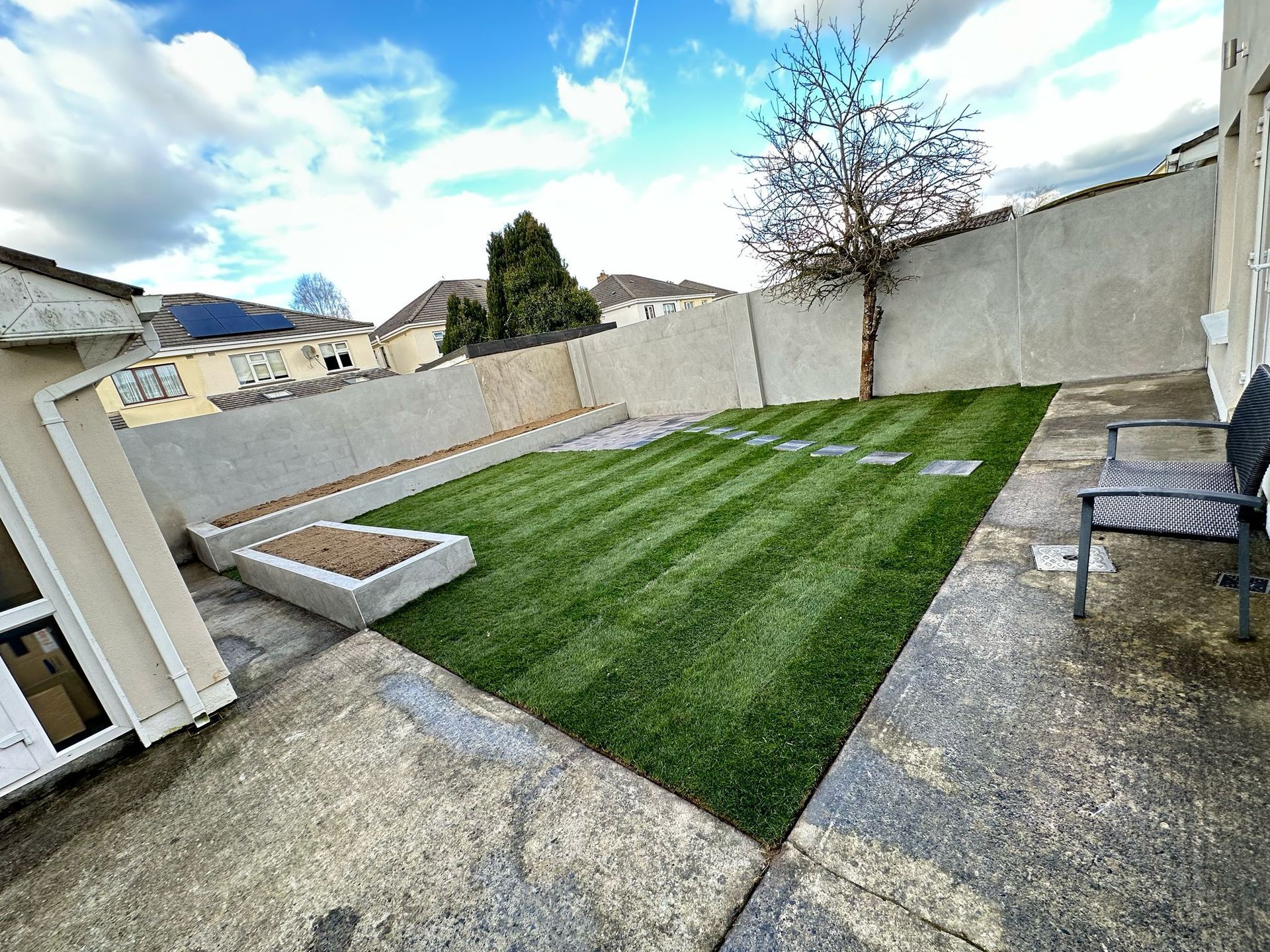 Backyard with striped green lawn, concrete patio, and gray walls under a cloudy sky.