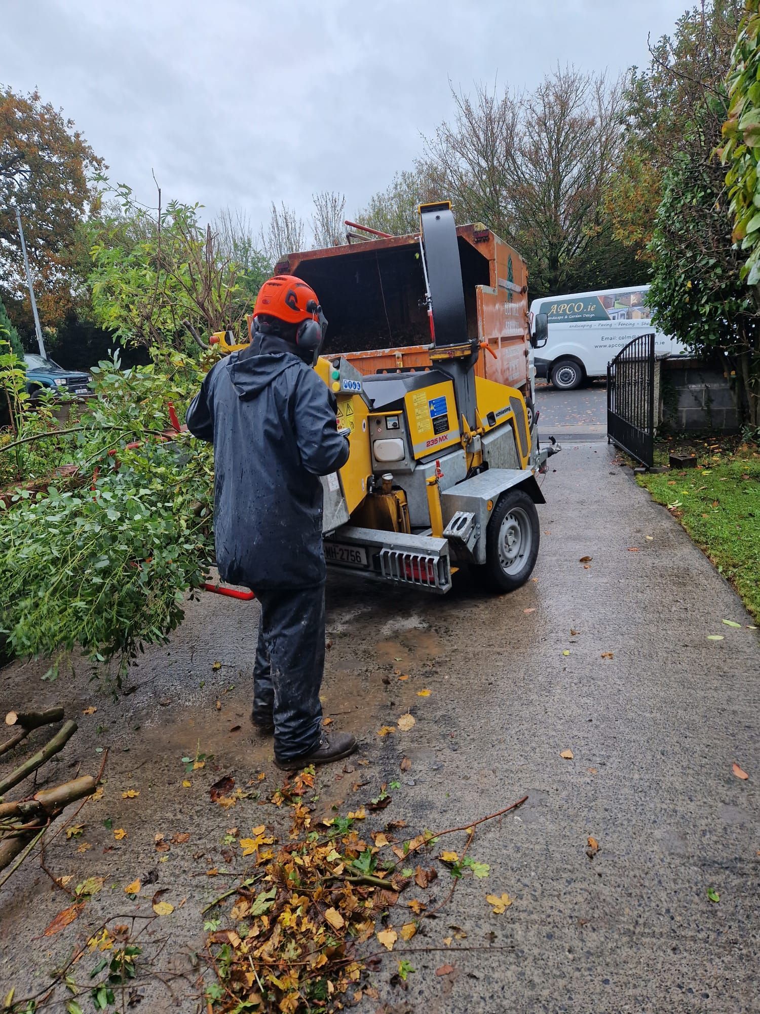 Person feeding branches into a wood chipper on a driveway; orange and yellow machine, cloudy day.