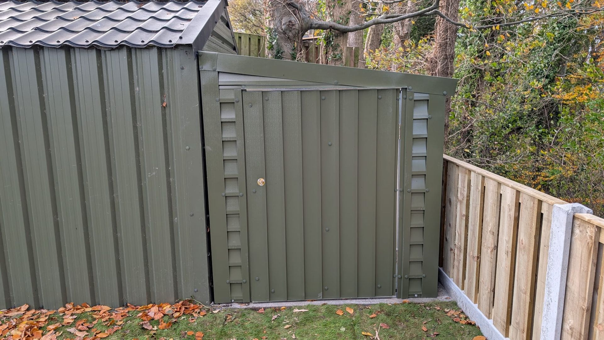 Green shed with door, next to wooden fence. Autumn leaves on the ground.