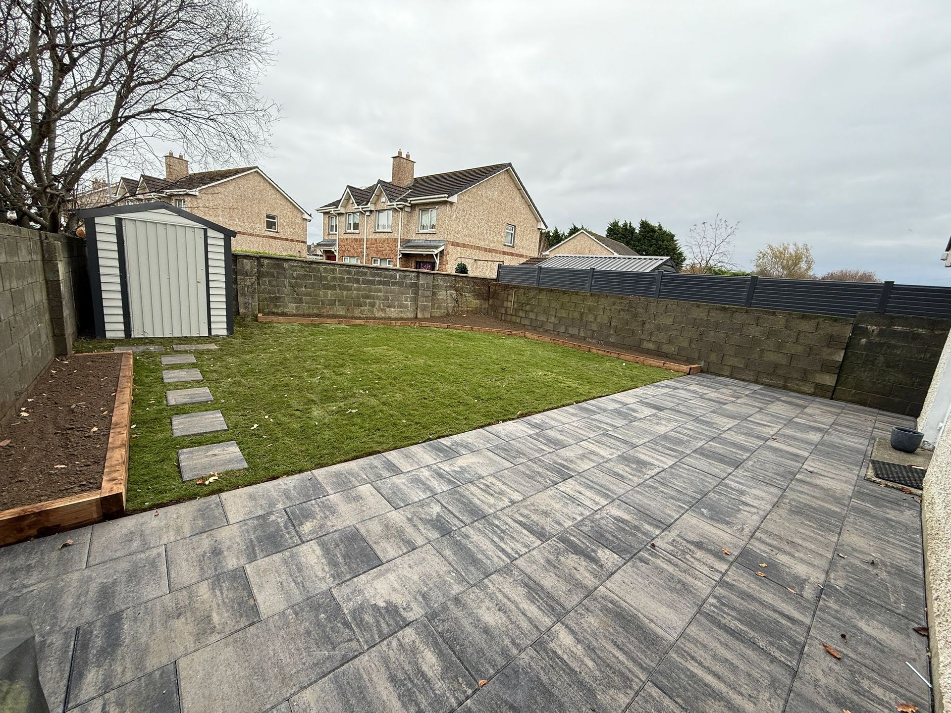 Backyard with lawn, patio, shed, and stone walls under overcast sky.