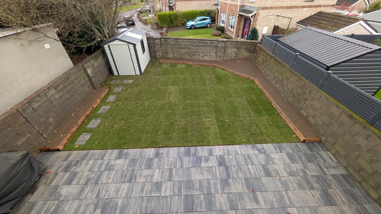 Overhead view of a small backyard: grass lawn, gray brick patio, shed, and brick border.