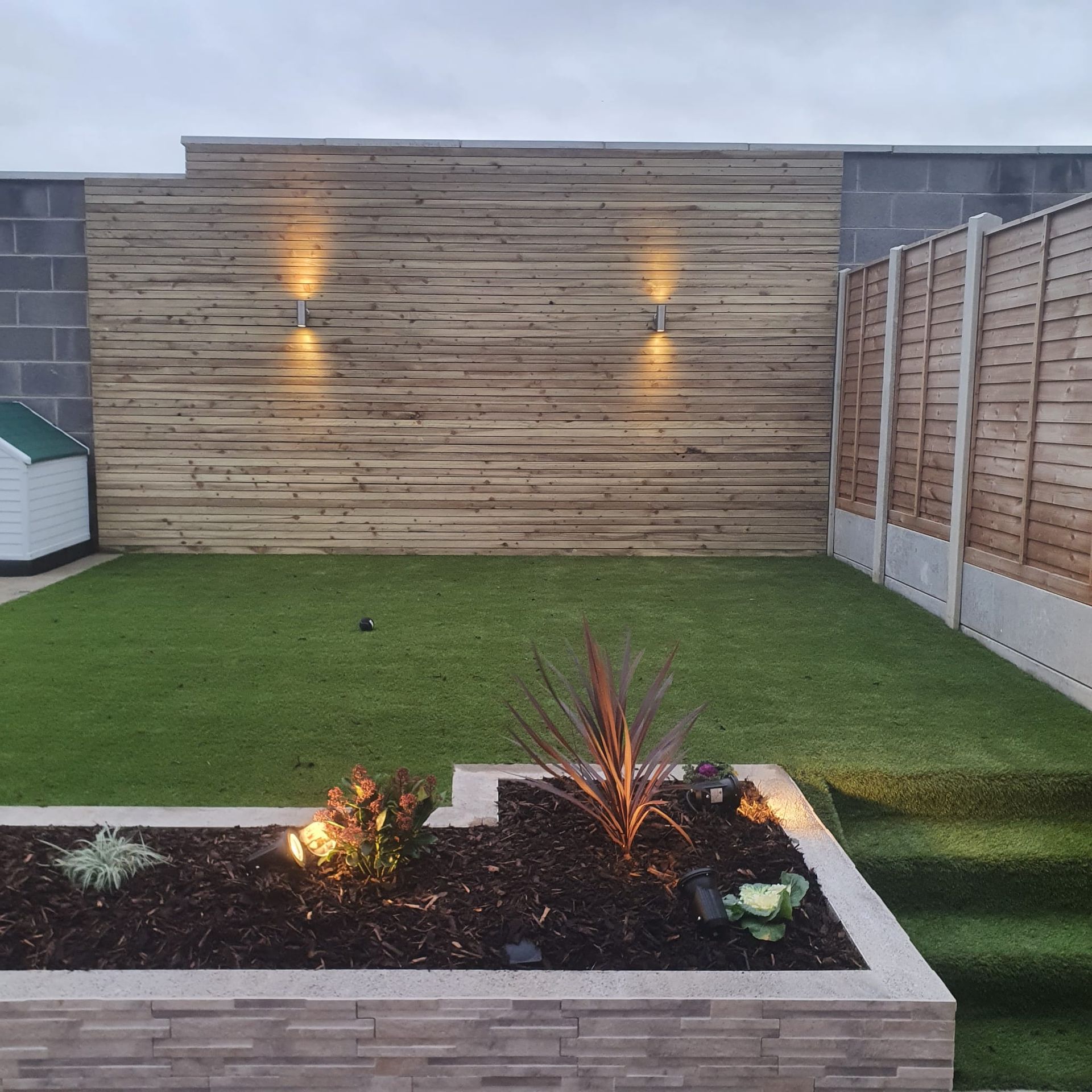 A backyard with a wooden fence, lawn, and raised garden bed. Two wall lights illuminate the wood.