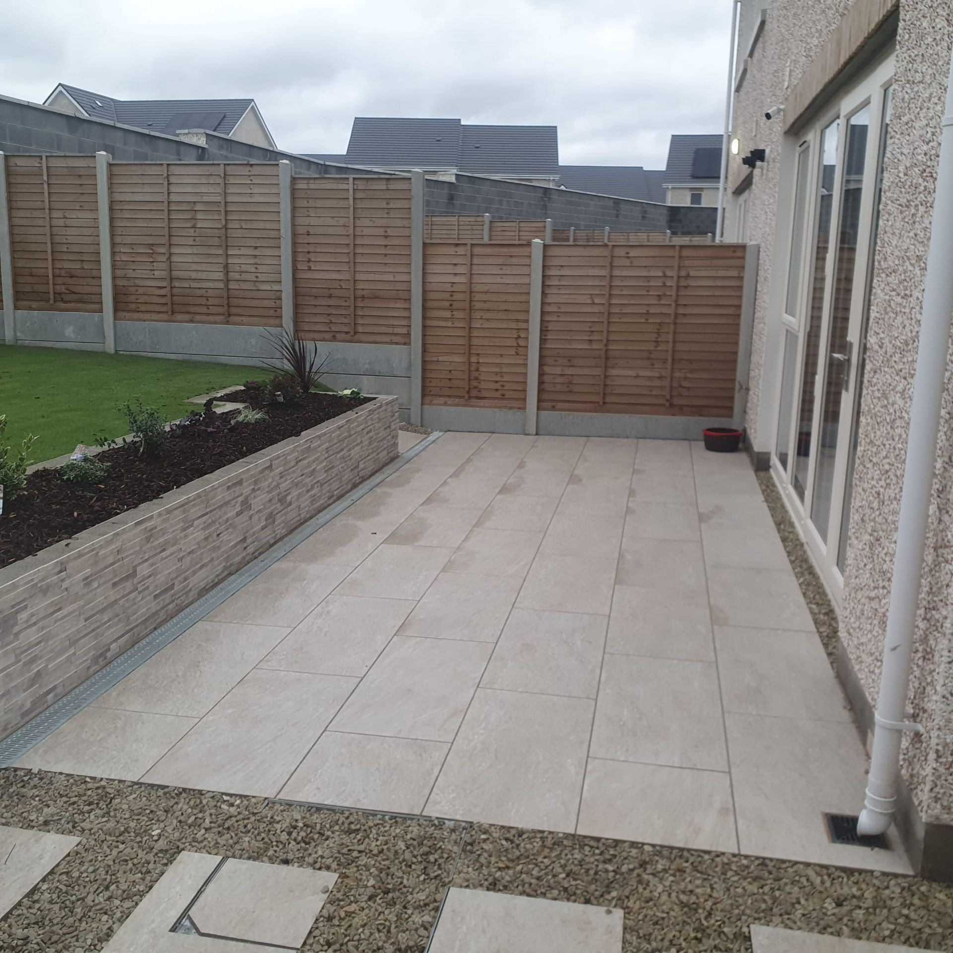 Patio with light-colored tiles, bordering a lawn and raised garden bed, next to a house with a fence in the background.