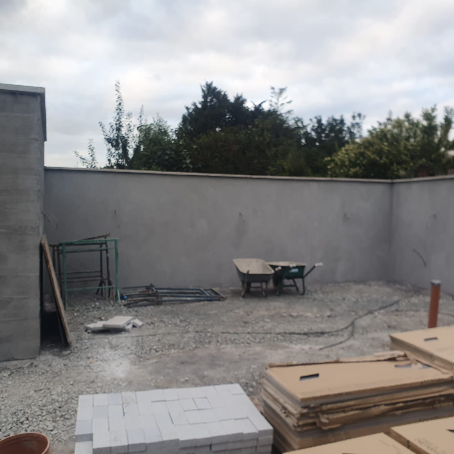 A concrete walled outdoor area under construction, with tools, materials, and a wheelbarrow present. Overcast sky.