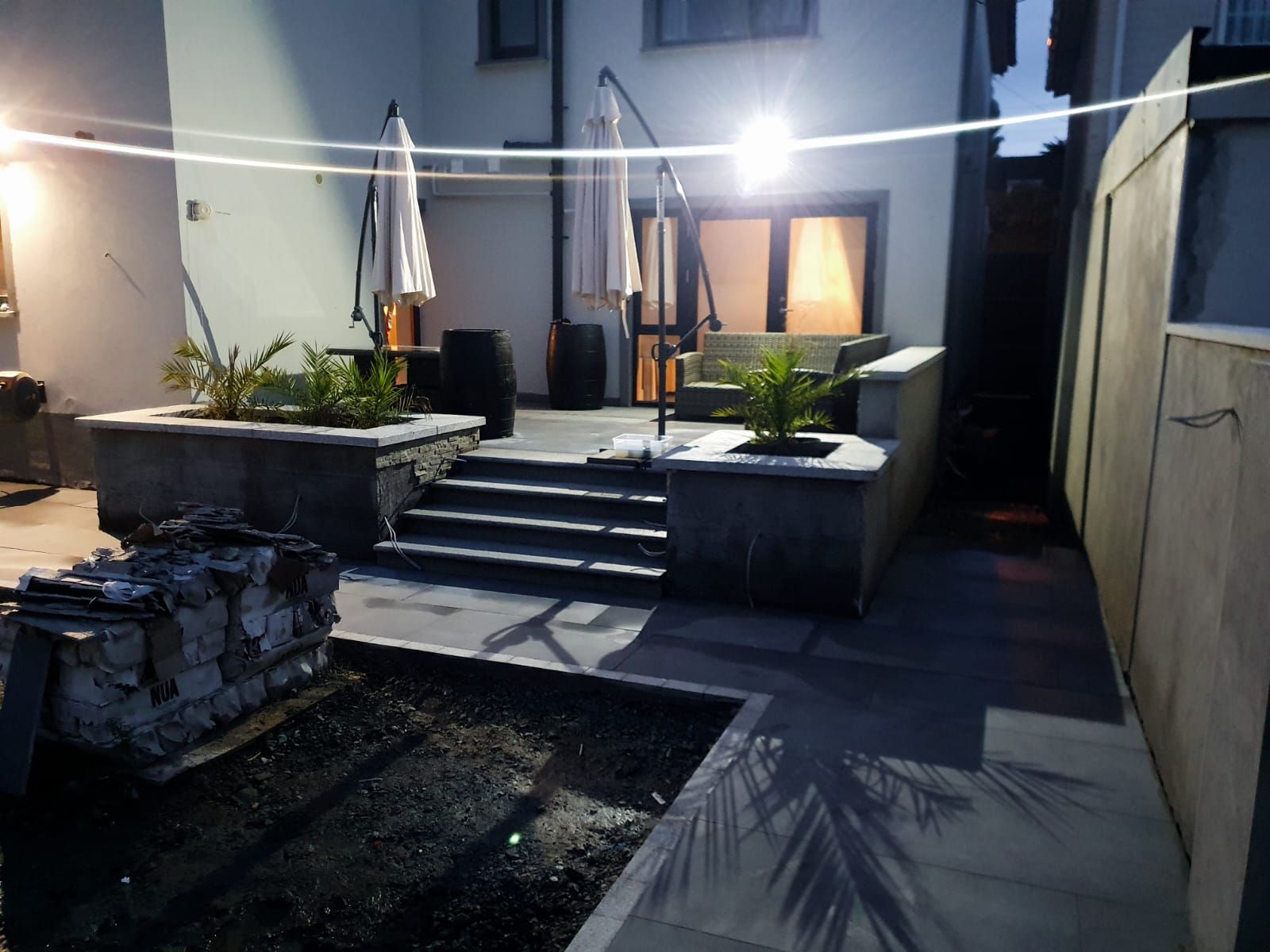 Backyard patio with steps, planters, and umbrellas, illuminated by overhead lights at dusk.