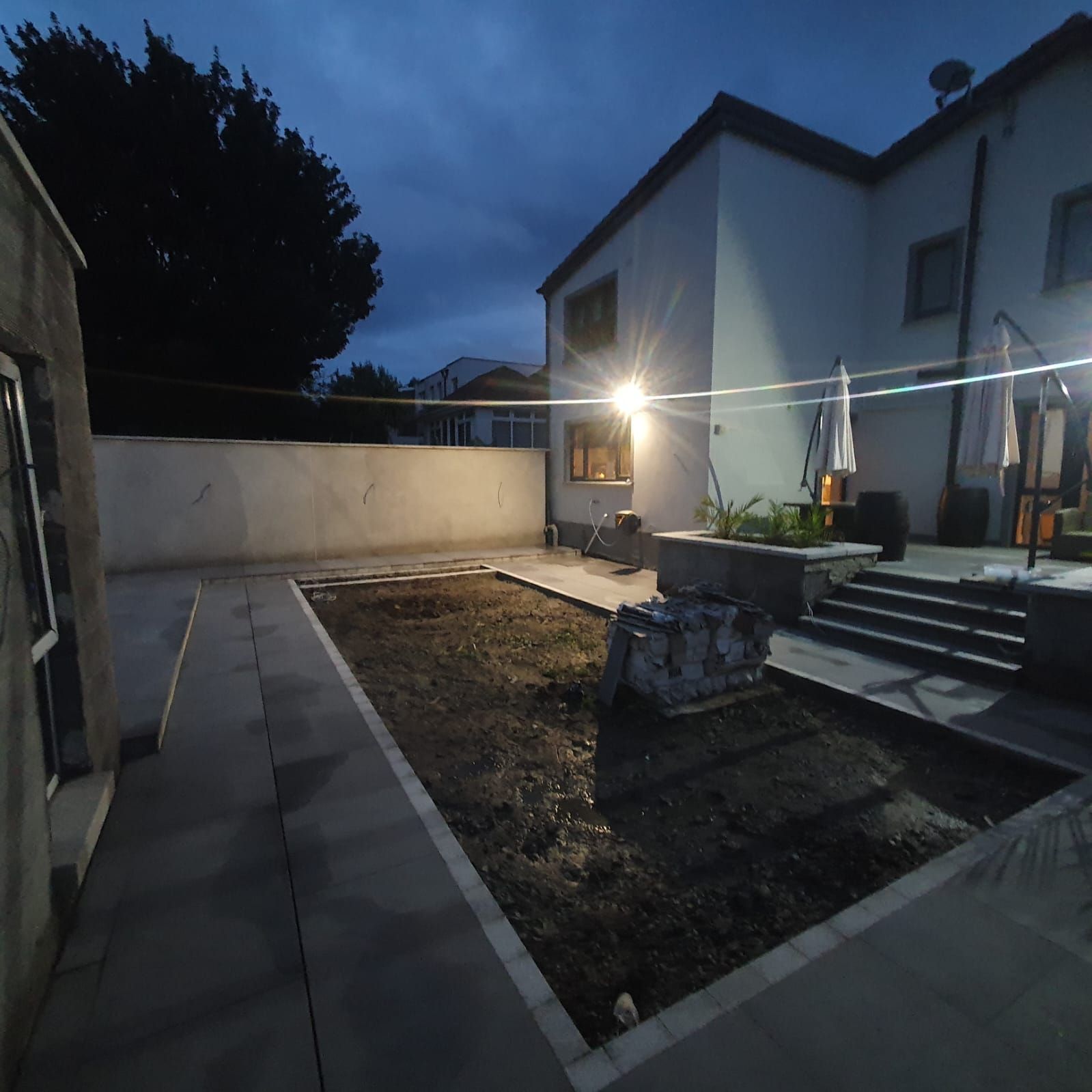 Backyard at dusk with dirt area, concrete, and house with lights.