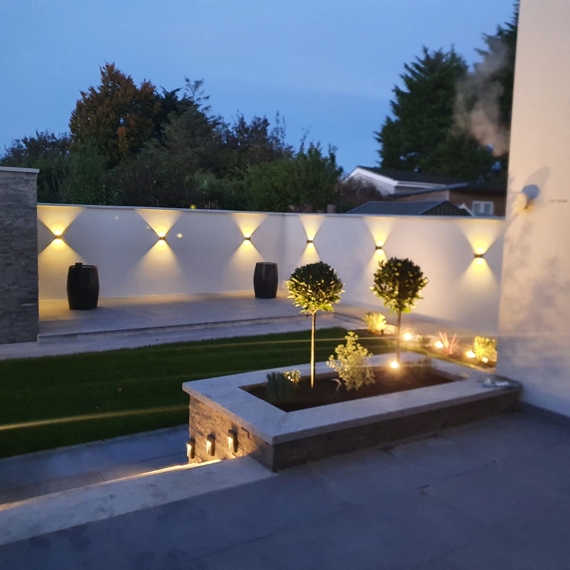 A modern outdoor garden at dusk with up-lighting on a white wall, a raised flower bed, and manicured trees.