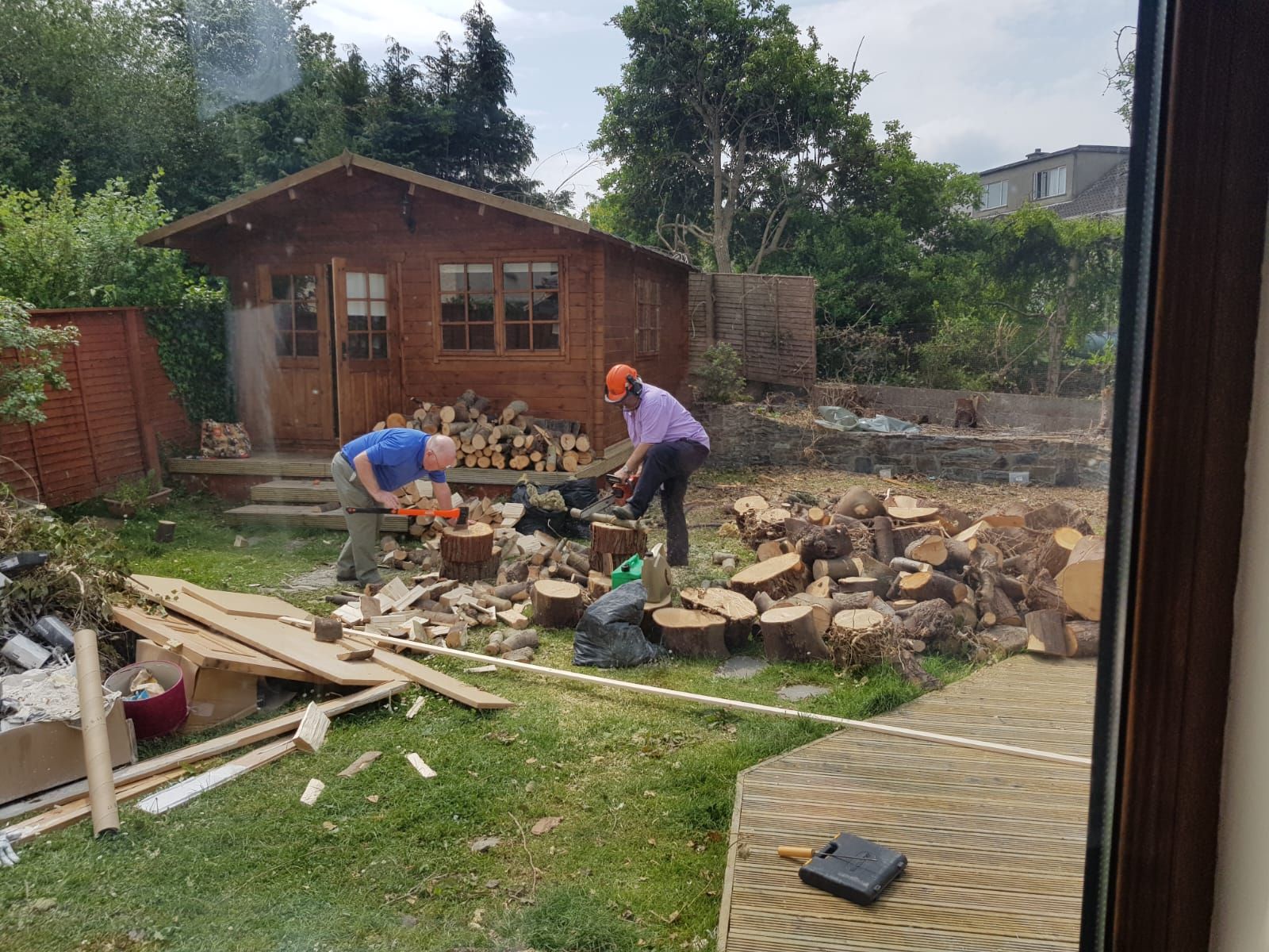 Two people cutting wood in a backyard with a shed. One wears an orange helmet, the other a blue shirt.
