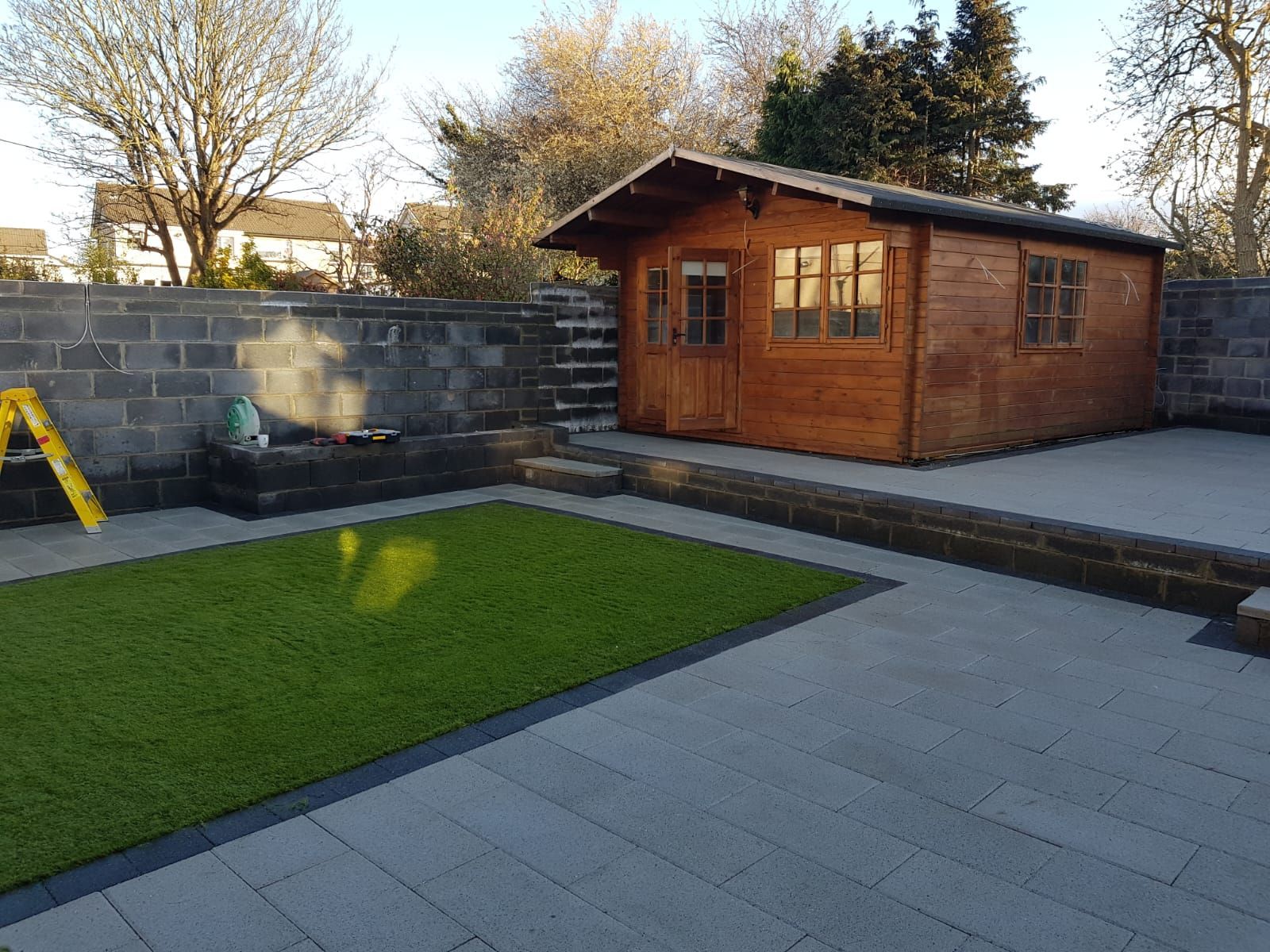 Wooden shed with windows in a landscaped garden with artificial green grass and stone patio.