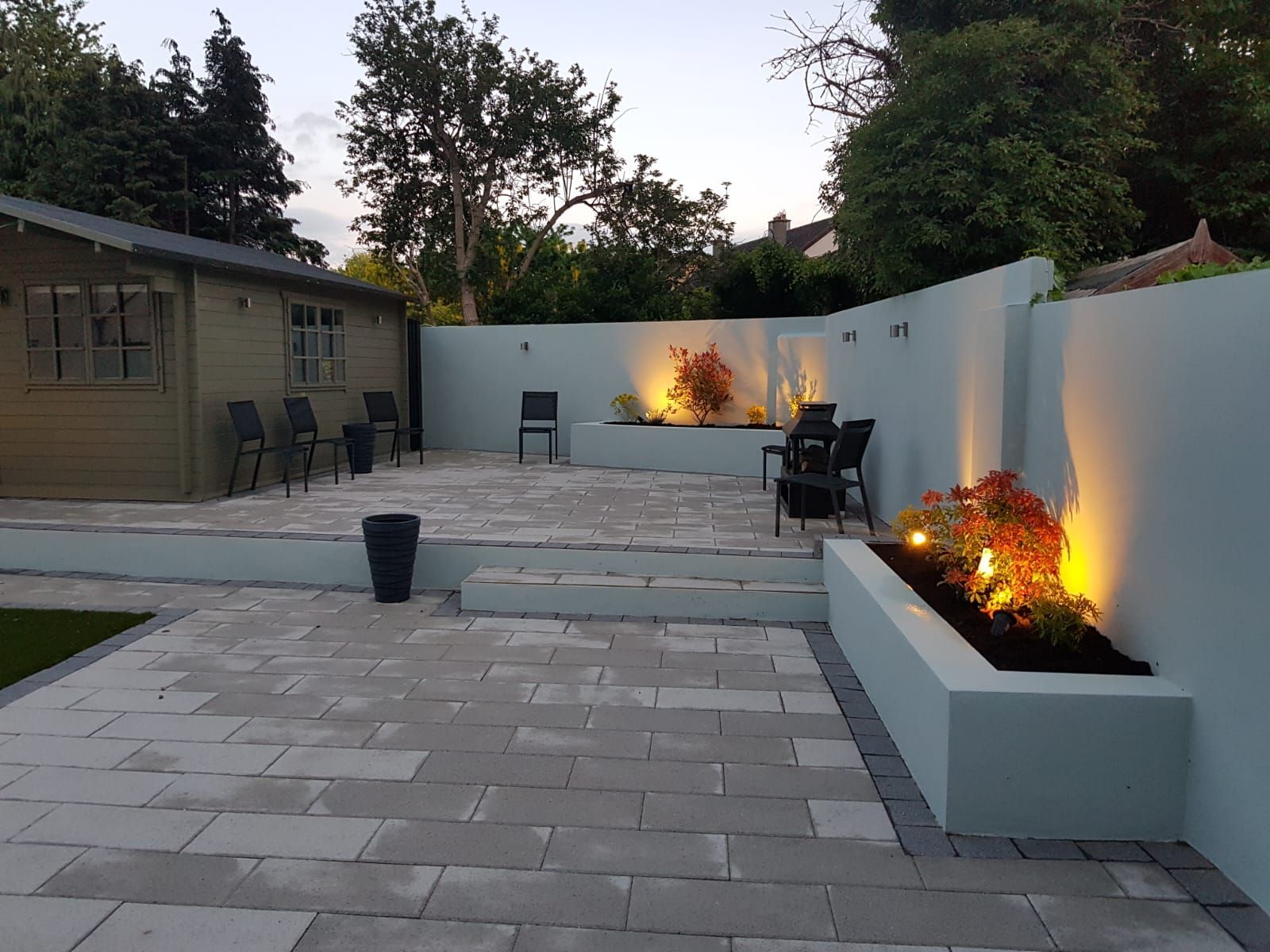 A paved patio with light blue walls, plants, and seating near a wooden shed at dusk.
