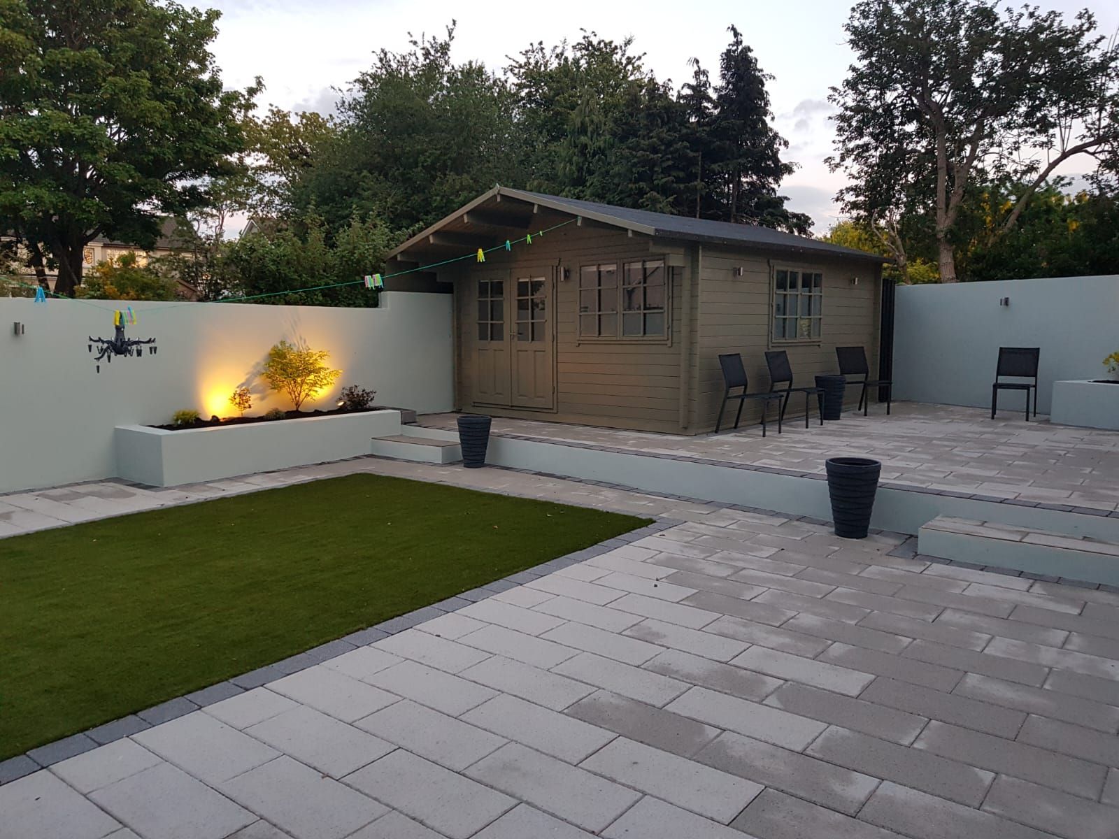 Backyard with gray patio, green lawn, shed, and light-lit retaining wall, plants, and chairs.