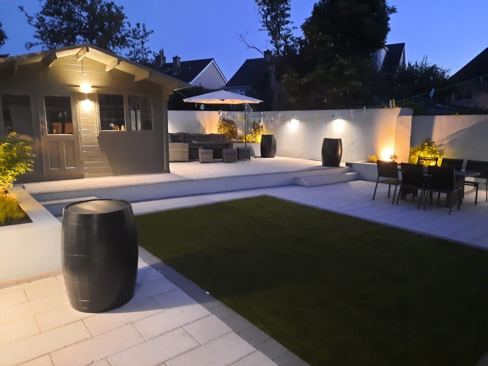 Outdoor patio at dusk with illuminated features, including a shed, seating area, and artificial lawn.