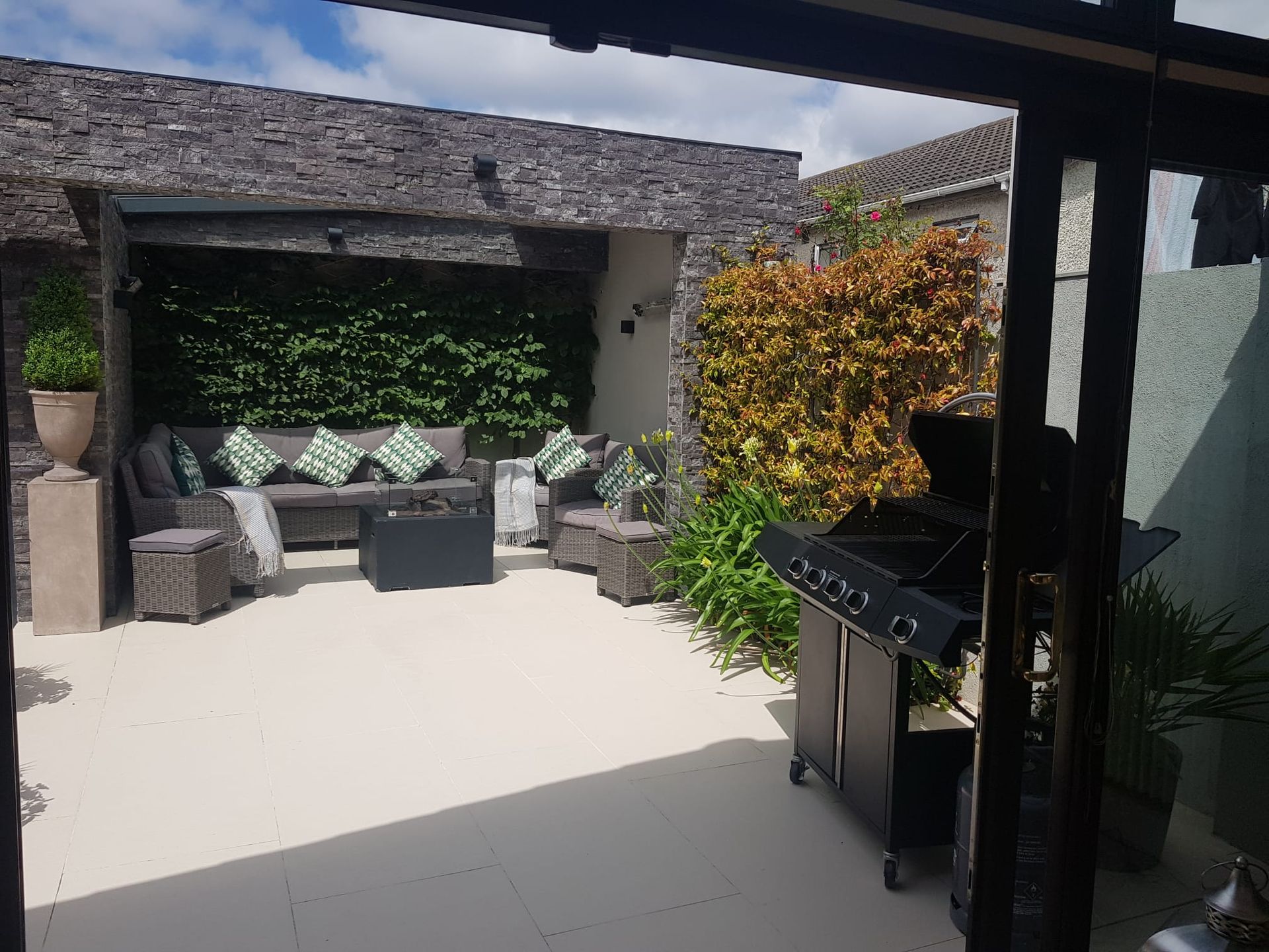 Outdoor patio with stone wall, seating area, and grill. Green and orange foliage.