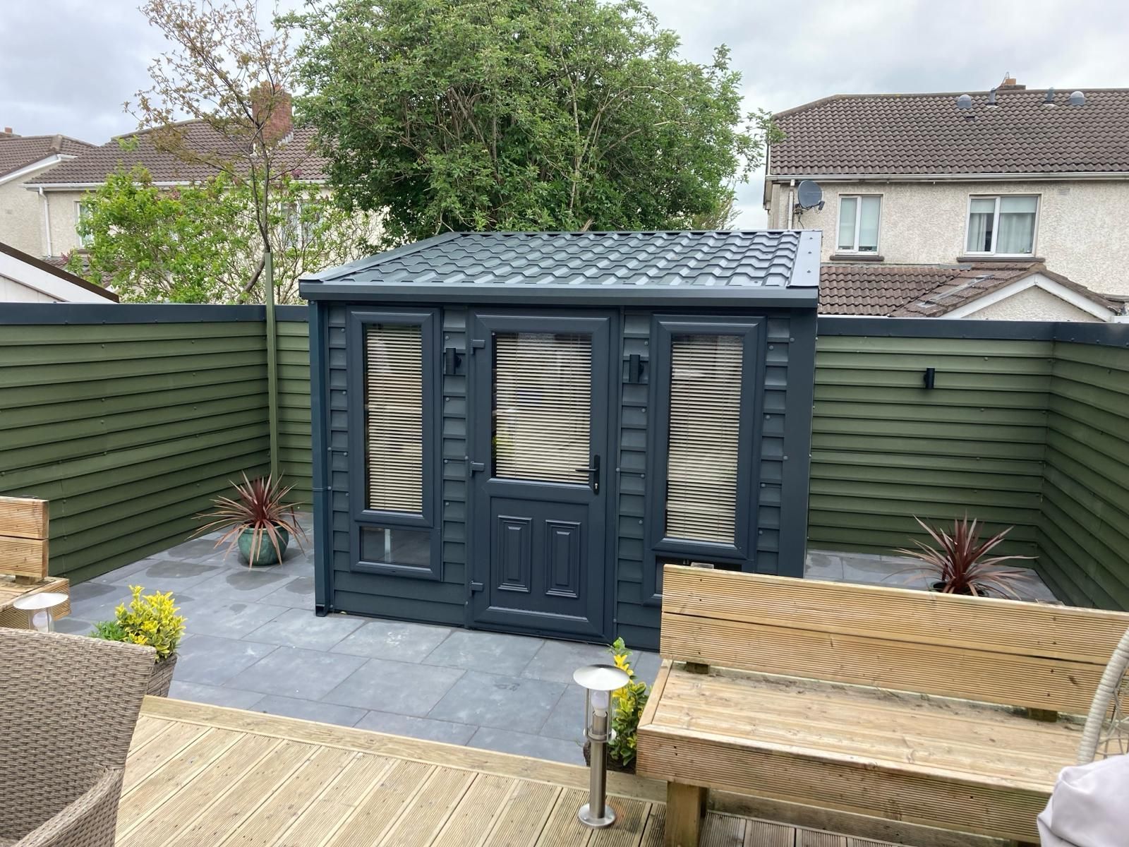 Dark blue shed with shuttered windows in a backyard with a gray tiled patio and green horizontal fence.
