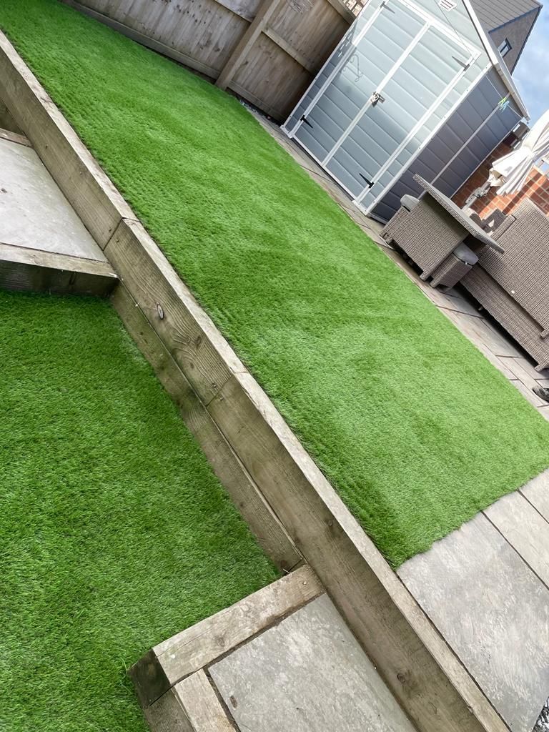 Artificial green lawn bordered by wood, with gray patio and shed in the background.