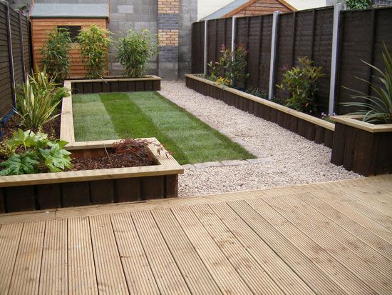 Wooden deck leads to gravel path beside raised garden beds with grass and plants.