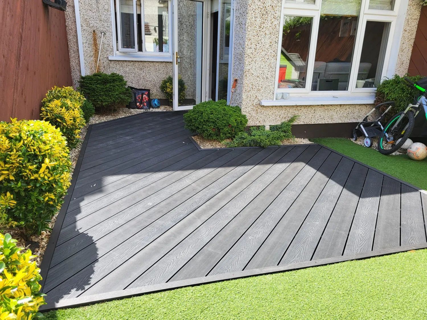 Backyard with dark gray composite deck surrounded by green turf and shrubs, with house in background.