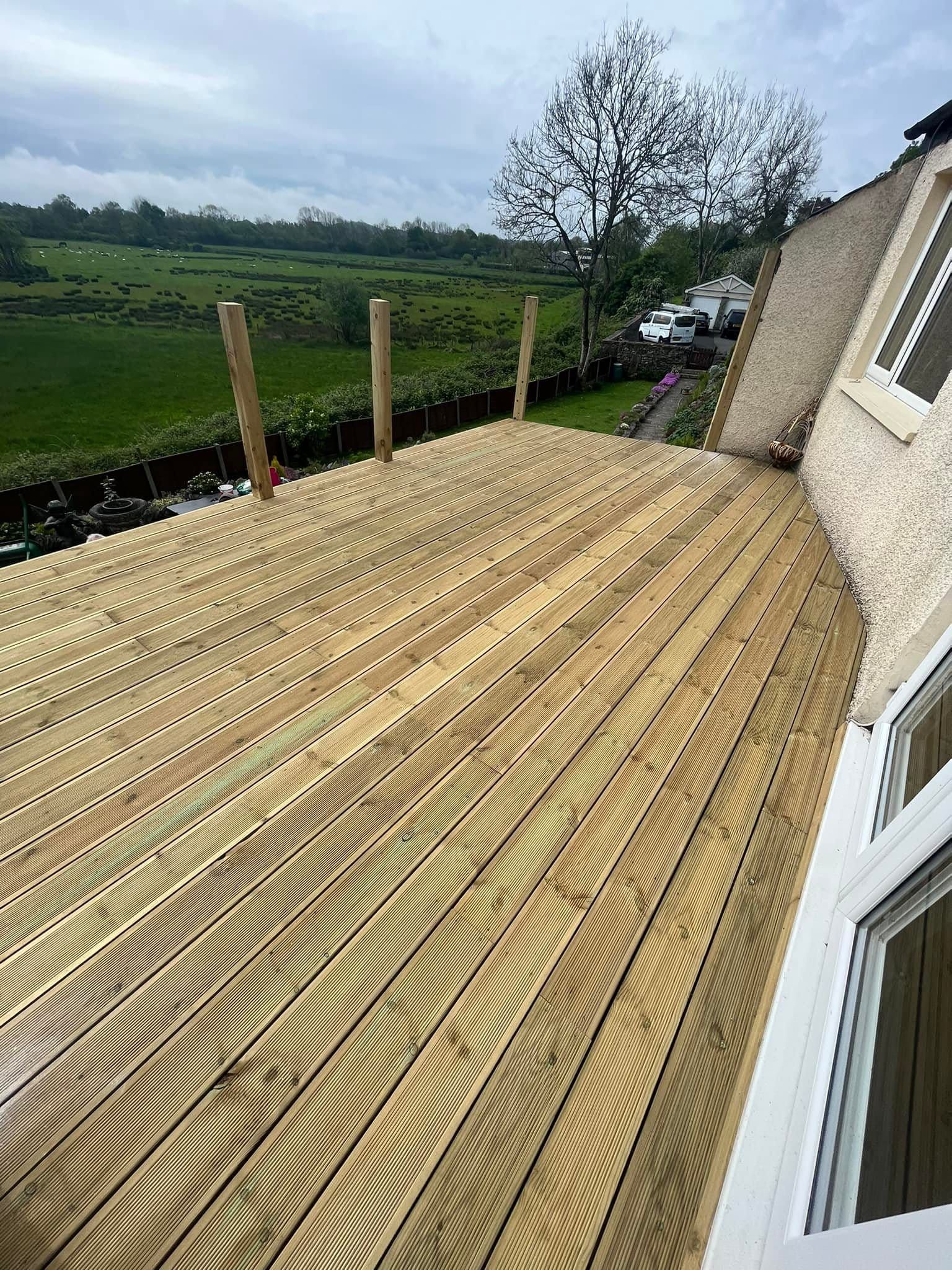 Wooden deck next to a building, overlooking a green landscape with posts and a tree.