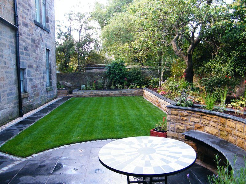 A backyard with a stone wall, lawn, patio, and table. Trees and foliage in the background.