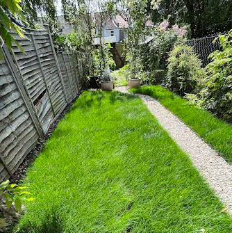 Narrow backyard with gravel path, bordered by lush green grass and a wooden fence.