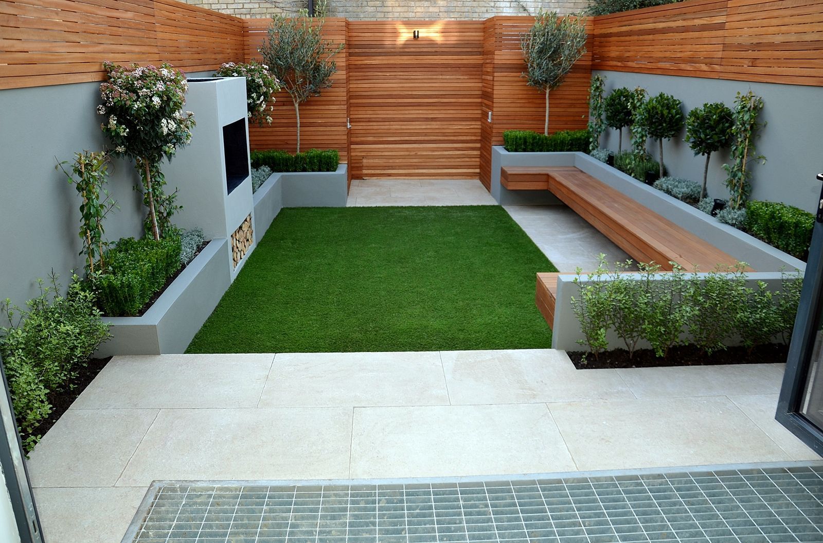 Modern backyard with green turf, built-in wooden bench, fireplace, and raised garden beds along perimeter.