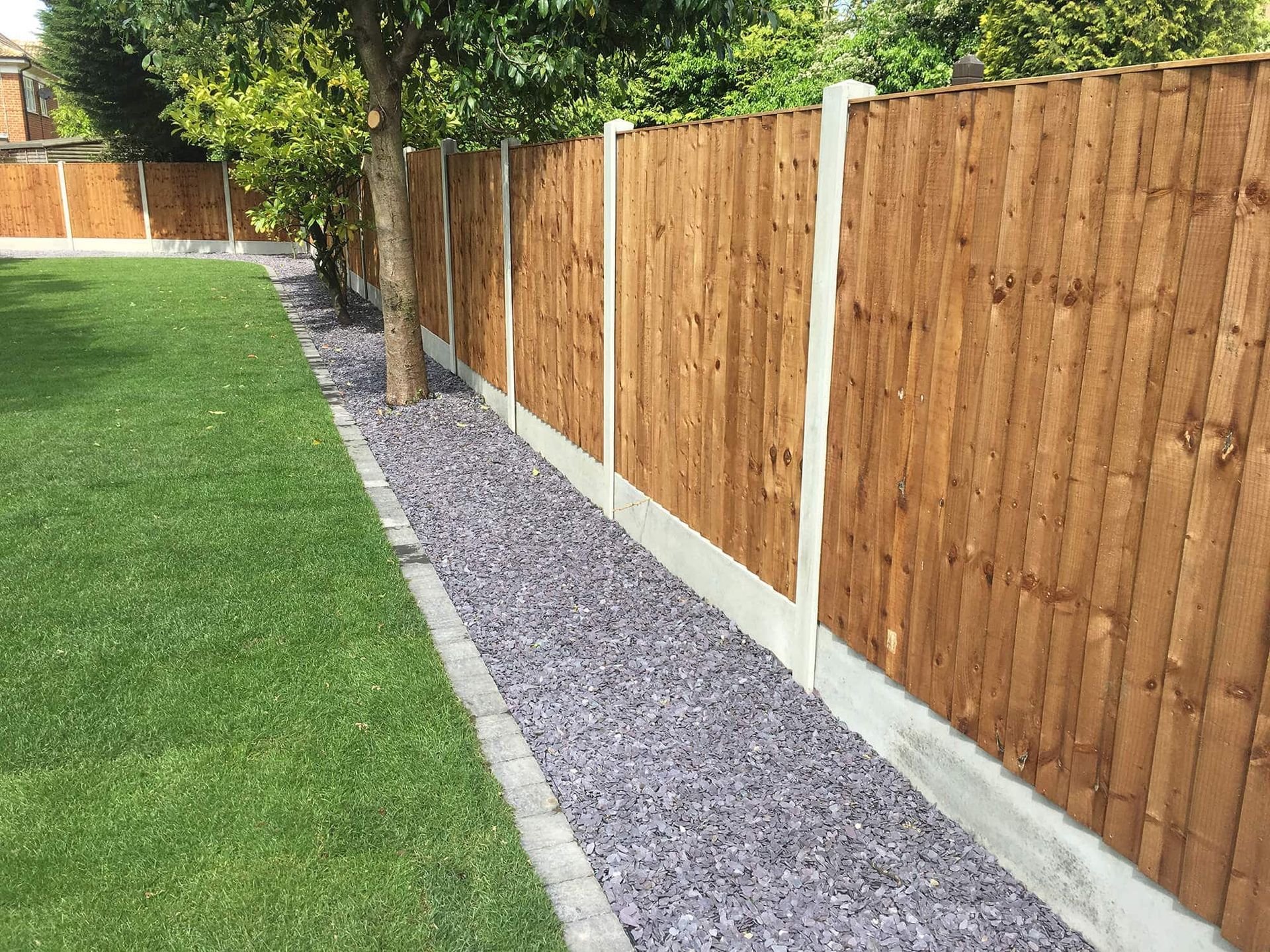 Wooden fence with concrete posts and base next to a gravel bed bordering a grassy lawn.