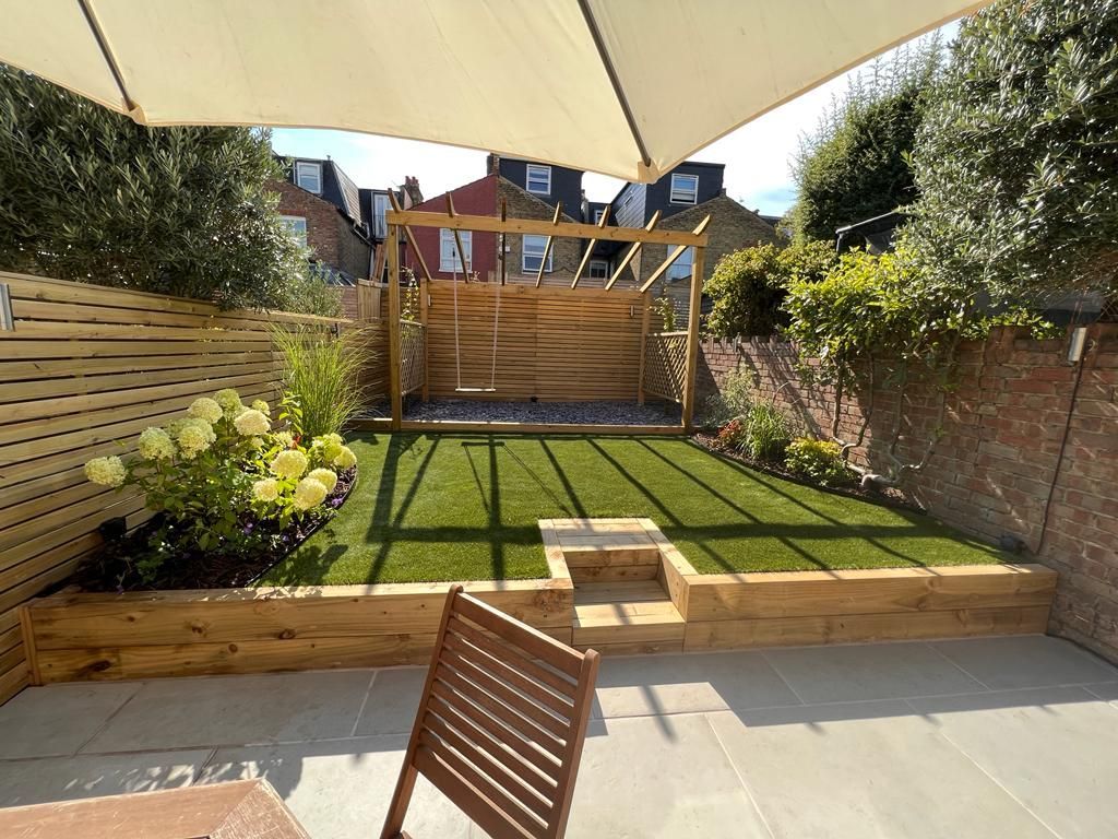 Backyard patio with wooden fence, artificial grass, and garden with a structure and a patio umbrella.