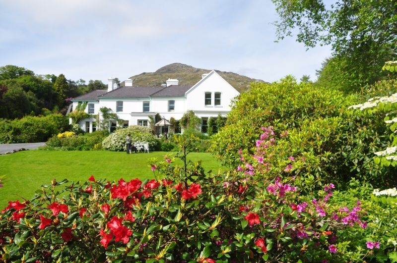 White house with green lawn and colorful flowers, mountains in the background.