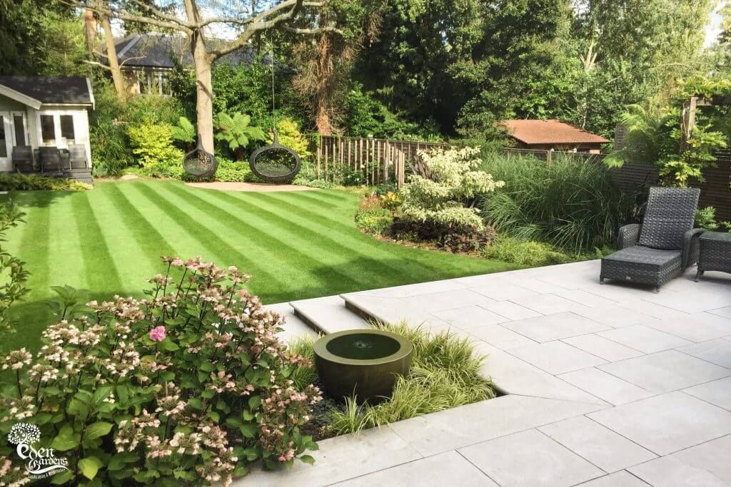 Neat lawn with striped mowing pattern, patio, and garden features: shrubs, seating, and small structures.