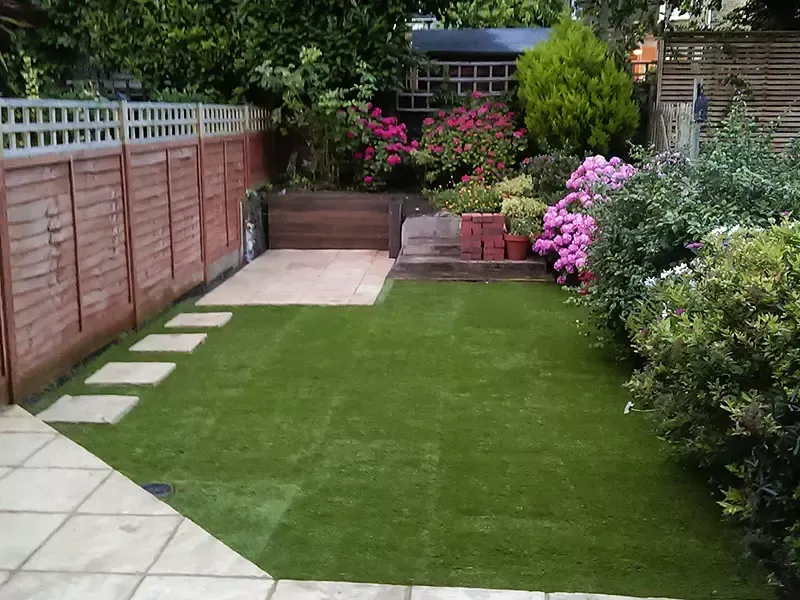 A lush green backyard with a paved patio and a wooden fence, flowers, and shrubbery.