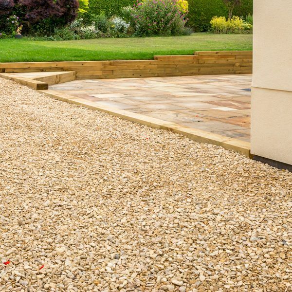 Gravel pathway borders a flagstone patio with wooden retaining walls, leading to a lawn with greenery.