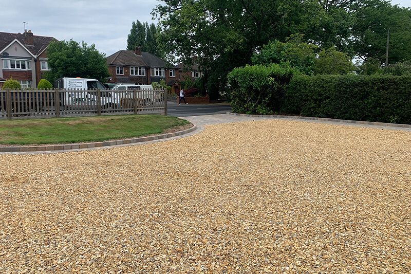 Gravel driveway in front of houses; green grass, low wooden fence, and hedge separate driveway from road.