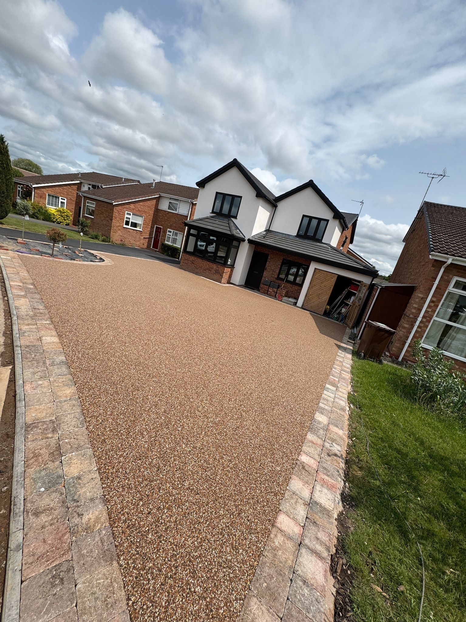 Gravel driveway leading to a white house with black trim. Brick border and blue sky with clouds.
